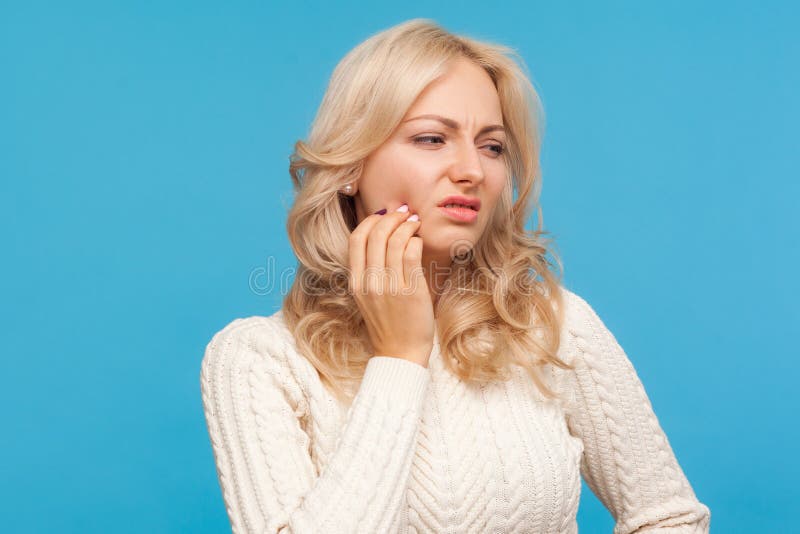 Closeup upset blond woman grimacing touching cheek, feeling unpleasant acute tooth pain, gums inflammation, dentistry. Indoor studio shot isolated on blue stock image