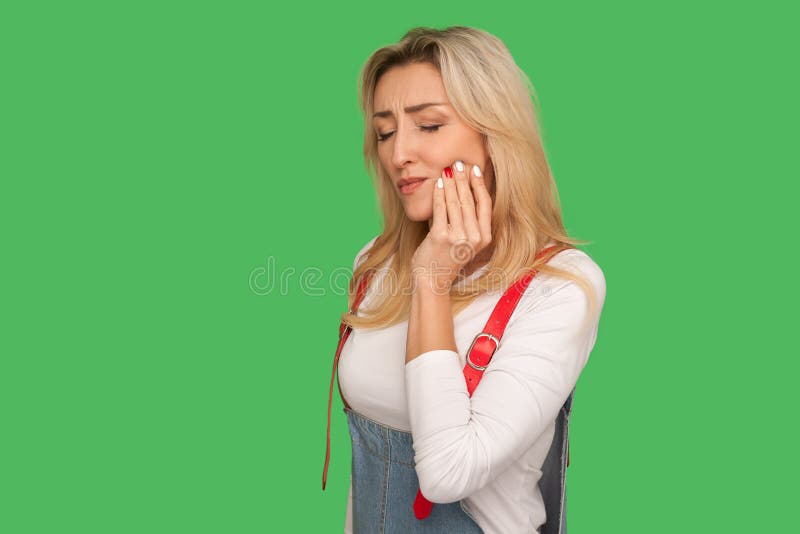 Dental problems, toothache. Portrait of sick adult woman in denim overalls touching sore cheek, suffering from cavities. Cracked teeth, gum recession. indoor stock photography