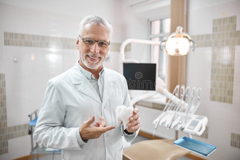 Joyful dentist holding a big molar tooth model. Cheerful elderly dental specialist holding and pointing to a big white tooth replica while explaining teeth royalty free stock photos