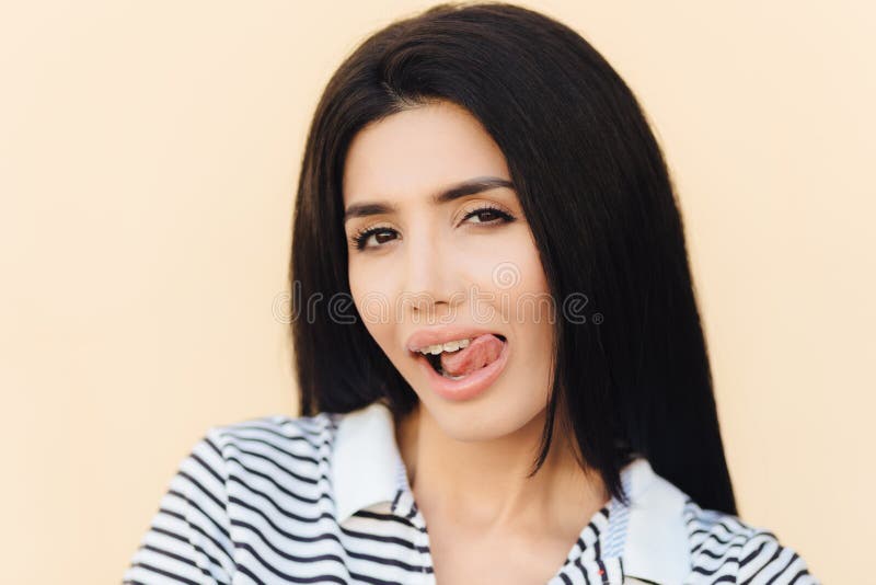 Portrait of attractive female model licks mouth with tongue, has curious look, healthy skin and dark hair, cunning expression, iso stock photos